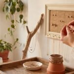 Crafter's hands arranging jewelry on a DIY picture frame earring holder on a white wall beside a driftwood display and clay ring dish.