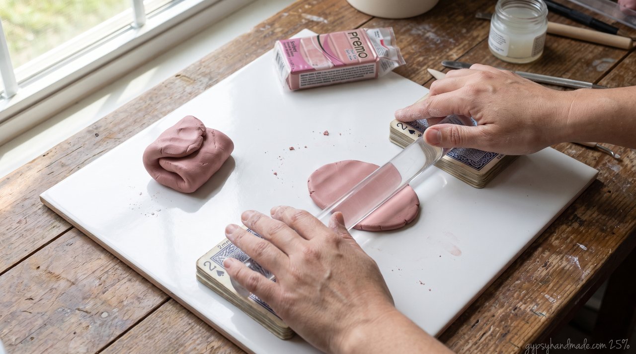 Close-up of hands rolling pink polymer clay on a ceramic tile with playing card thickness guides for consistent 2mm depth.