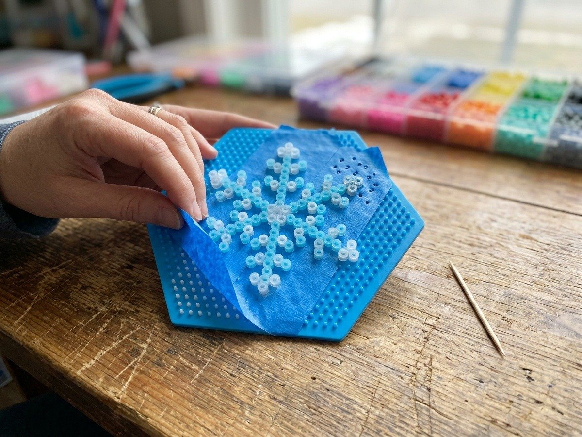 Demonstration of blue painter tape method applied over arranged Perler bead snowflake to protect plastic pegboard from heat